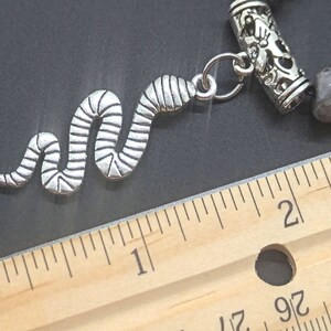 May include: A silver snake charm with a textured design, attached to a silver bead bracelet with a grey and white stone bead. The bracelet is measured against a wooden ruler.