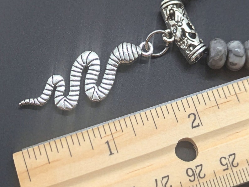 May include: A silver snake charm with a textured design, attached to a silver bead bracelet with a grey and white stone bead. The bracelet is measured against a wooden ruler.