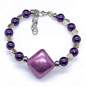 May include: A purple and silver bracelet with a large, iridescent, diamond-shaped bead in the center. The bracelet is made with small, round, purple beads and silver spacer beads.
