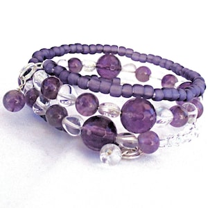 May include: A multi-strand bracelet with purple and clear beads. The bracelet is made with small, round beads and larger, round beads. The bracelet has a silver charm attached to it.