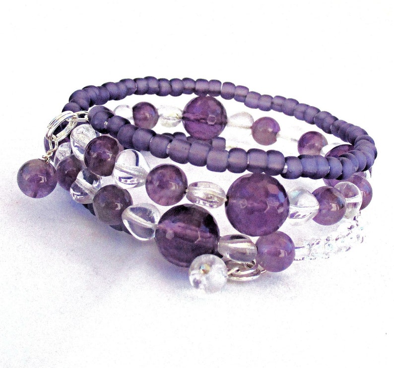 May include: A multi-strand bracelet with purple and clear beads. The bracelet is made with small, round beads and larger, round beads. The bracelet has a silver charm attached to it.