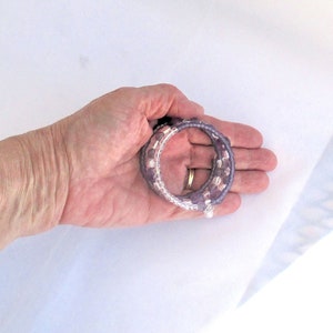 May include: A set of three purple and clear beaded bracelets with a silver charm. The bracelets are stacked on top of each other and are held in a hand.