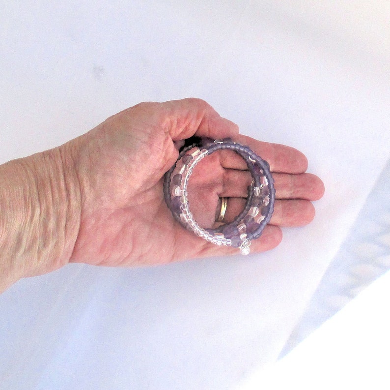May include: A set of three purple and clear beaded bracelets with a silver charm. The bracelets are stacked on top of each other and are held in a hand.