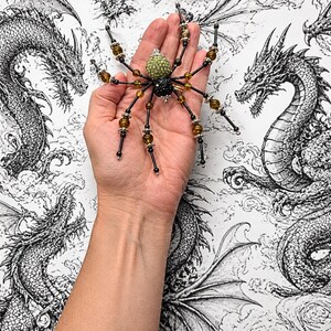 May include: A decorative spider ornament featuring a textured light green head and a black body, with eight long legs adorned with alternating black and amber-colored beads separated by silver-toned metal spacers and small silver end caps.