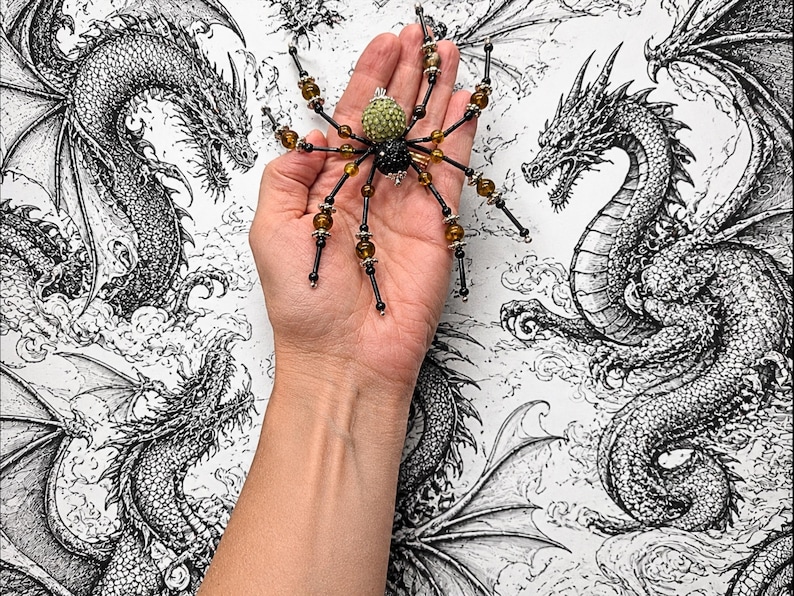 May include: A decorative spider ornament featuring a textured light green head and a black body, with eight long legs adorned with alternating black and amber-colored beads separated by silver-toned metal spacers and small silver end caps.