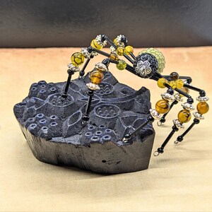 May include: A decorative spider figurine crafted from black wire, amber-colored crackle beads, and silver-toned metal accents, featuring a textured green bead body, perched atop a dark, carved, irregular block with a floral relief pattern.