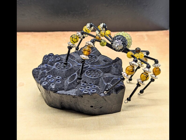 May include: A decorative spider figurine crafted from black wire, amber-colored crackle beads, and silver-toned metal accents, featuring a textured green bead body, perched atop a dark, carved, irregular block with a floral relief pattern.