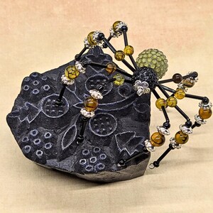 May include: A decorative spider figurine crafted from black wire, featuring amber-colored crackle beads and silver-toned spacers on its legs. The spider has a black beaded head and a textured, light green spherical body, perched on a black block.