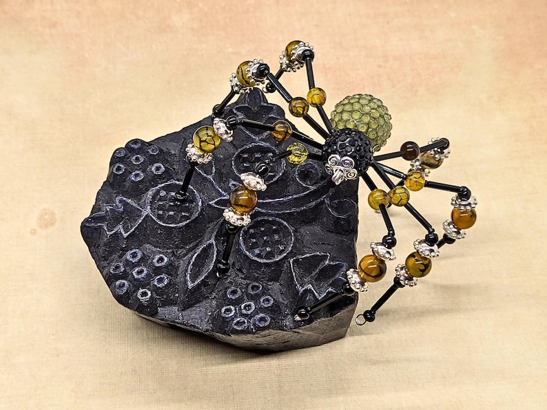 May include: A decorative spider figurine crafted from black wire, featuring amber-colored crackle beads and silver-toned spacers on its legs. The spider has a black beaded head and a textured, light green spherical body, perched on a black block.