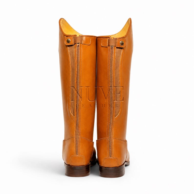 May include: A pair of tall, tan leather riding boots. The boots have a zipper up the back and a small strap at the top. The brand name "NUVE LEATHER" is embossed on the side of the boots. The boots are set against a white background.