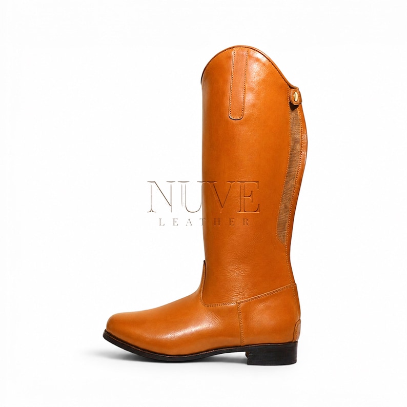 May include: A tall, equestrian-style riding boot in a warm, tan color. The leather boot features a rounded toe, a tall shaft, and a decorative strap near the top. The brand "NUVE LEATHER" is printed on the side.