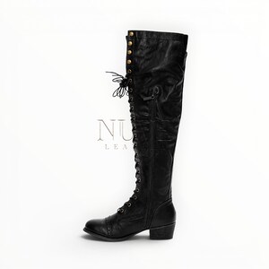 May include: Black leather knee-high boot with lace-up front and side zipper. The boot features a small heel and decorative gold-colored studs near the top. The brand name "NU LEATHER" is visible in the image.
