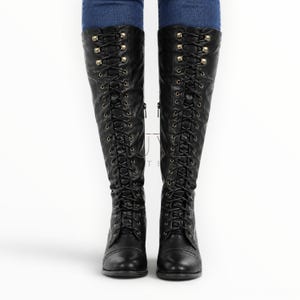 May include: Black knee-high boots with a lace-up design and gold-colored eyelets. The boots have a round toe and a zipper on the side. The boots are made of a smooth material and are worn with blue jeans.