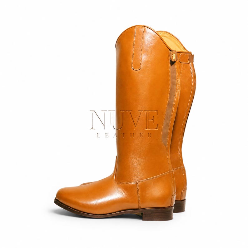 May include: A pair of tall, tan leather riding boots. The boots have a classic design with a rounded toe, a tall shaft, and a small heel. The boots are made of smooth, polished leather and feature a strap and buckle detail near the top. The brand name "NUVE LEATHER" is visible.