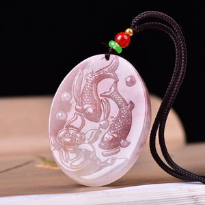 Peut inclure: Un pendentif ovale rose clair orné d'une sculpture en relief représentant deux poissons koï et une fleur de lotus. Le pendentif est suspendu à un cordon noir agrémenté d'une petite perle verte et d'une perle rouge avec des embouts dorés.