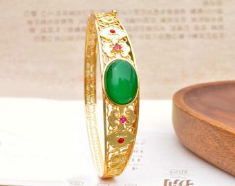 May include: Gold-toned bangle bracelet featuring an oval, vibrant green gemstone at the center. The bracelet has an openwork design with floral and red gemstone accents. The bracelet is displayed on a white surface.