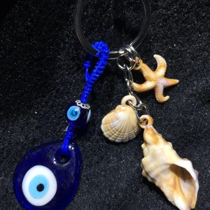 以下が含まれることがあります： 青い邪視チャーム、ヒトデ、2つの貝殻が付いたシルバーのキーホルダー。邪視チャームは濃い青色で、白と黒の中心があります。貝殻はベージュ色で、ヒトデは明るいオレンジ色です。