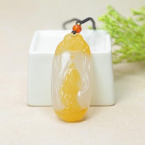 Op de afbeelding: Een gele en witte hanger met een gebeeldhouwde figuur. De hanger is opgehangen aan een zwart koord met een kleine oranje kraal. De hanger heeft een gedetailleerde gravure van een persoon in vloeiende gewaden.
