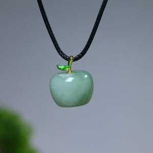 May include: A light green apple pendant necklace. The apple-shaped pendant has a glossy finish and is accented with a gold-colored stem and a green leaf. The necklace features a black cord.
