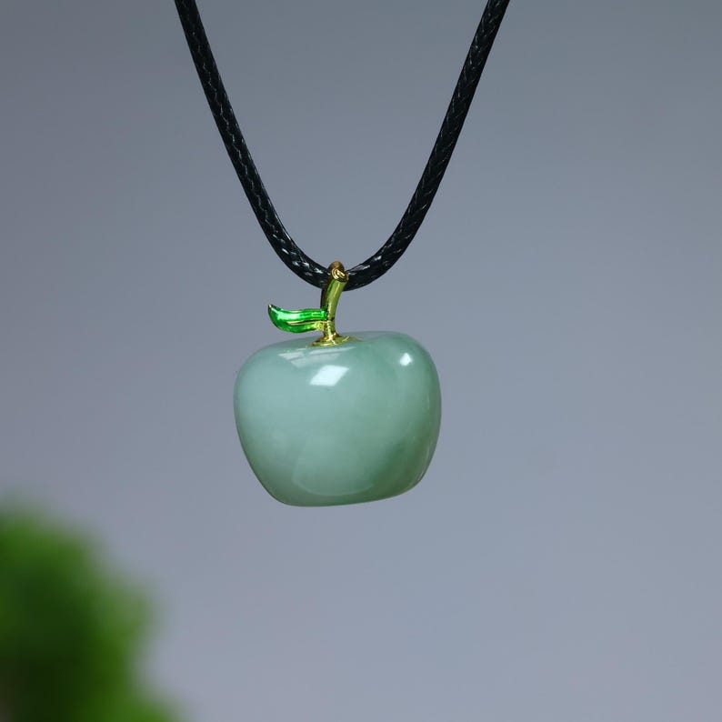 May include: A light green apple pendant necklace. The apple-shaped pendant has a glossy finish and is accented with a gold-colored stem and a green leaf. The necklace features a black cord.