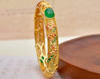 May include: A gold-colored bangle bracelet with an oval green stone and floral designs in red, pink, and green. The bracelet has an intricate pattern and is likely made of metal.