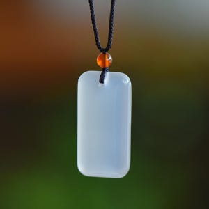 Puede incluir: Un collar con colgante rectangular de color claro con esquinas redondeadas. El colgante está suspendido de un cordón negro, con una pequeña cuenta naranja. El fondo es una mezcla borrosa de verde y marrón.