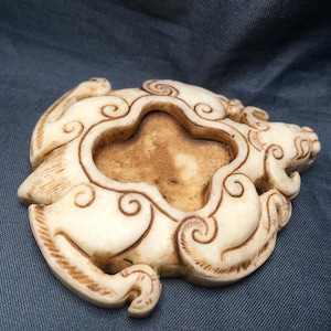 May include: Ivory-colored, carved decorative object with a central quatrefoil-shaped opening. The object features a stylized animal design with swirling cloud-like patterns. The piece has a weathered appearance, suggesting age or a vintage aesthetic.