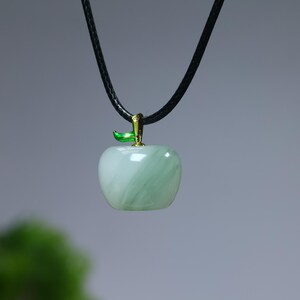 May include: A light green apple pendant with a black cord. The apple-shaped charm has a gold-colored stem and a green leaf. The pendant is made of a translucent material with a marbled effect.