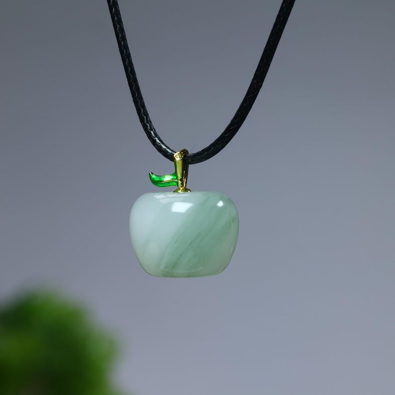 May include: A light green apple pendant with a black cord. The apple-shaped charm has a gold-colored stem and a green leaf. The pendant is made of a translucent material with a marbled effect.
