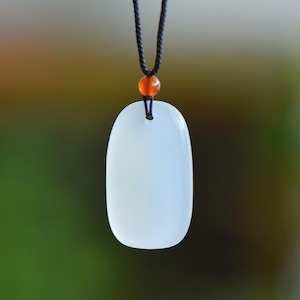 Puede incluir: Collar con colgante blanco de forma ovalada. El colgante liso y opaco está suspendido de un cordón negro. Una pequeña cuenta naranja se encuentra encima del colgante. El fondo es una mezcla borrosa de verde y marrón.