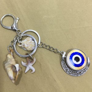 以下が含まれることがあります： 青と白の邪視チャーム、貝殻、ヒトデが付いたシルバーのキーホルダー。キーホルダーにはロブスタークラスプとチェーンが付いています。邪視チャームは丸く、シルバーの透かし模様の縁取りがあります。貝殻はベージュと白です。