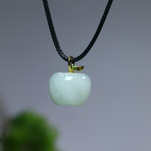 May include: A light green apple pendant necklace. The apple-shaped pendant has a glossy finish and is suspended from a black cord. The stem and attachment are gold-colored. The necklace is a jewelry item.
