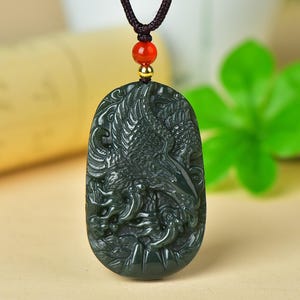 Può includere: Un ciondolo ovale verde scuro con un'intricata incisione in rilievo di un uccello con le ali spiegate. Il ciondolo pende da un cordino marrone scuro, accentuato da una piccola perlina color oro e una perlina arancione liscia e rotonda.