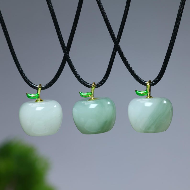 May include: Three apple-shaped pendants on black cords. The pendants are light green and white, with a gold-colored stem and a green leaf. The background is a soft gray.