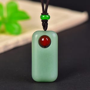 May include: A rectangular light green pendant with rounded corners featuring a circular cutout at the top containing a small, polished red bead. The pendant hangs from a dark brown cord accented with a single, small, bright green bead.