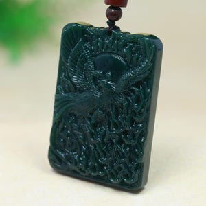 May include: A rectangular dark green jade pendant featuring an intricate relief carving of a phoenix bird with spread wings and flowing tail feathers. The pendant is suspended from a dark brown cord with a small matching bead at the top.