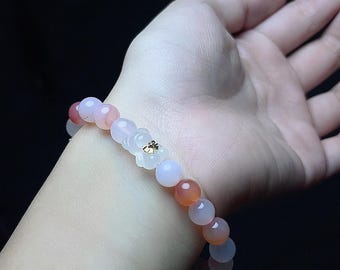 May include: A beaded bracelet featuring a mix of pink, white, and orange-toned round beads. A white flower-shaped charm with a small gold accent is centered on the bracelet. The bracelet is worn on a wrist.