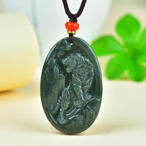 May include: Dark green jade pendant with a carved tiger design. The oval-shaped pendant is suspended from a brown cord with a red bead and gold-colored accents. The tiger is depicted in a detailed, seated pose.