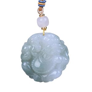 Peut inclure: Pendentif en jade vert clair représentant une figure sculptée, suspendu à un cordon tressé avec des accents bleus, dorés et beiges. Le pendentif est rond, avec des gravures détaillées et une finition polie.