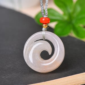 May include: A light pink pendant with a swirling design, suspended from a gray cord. A red bead and a gold-colored bead are positioned above the pendant. The pendant is made of a translucent material.