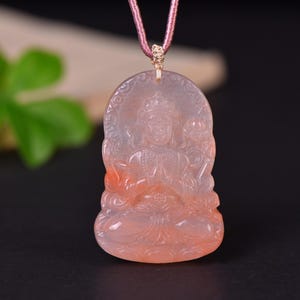 May include: A translucent, pale pink carved stone pendant featuring a seated deity figure with hands in a prayer position. The pendant has a smooth, rounded top edge and is suspended from a mauve-colored cord with a small, intricate knot detail.