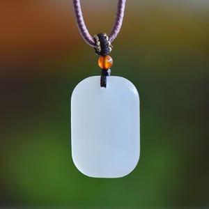 Puede incluir: Un collar con colgante rectangular blanco con bordes redondeados. El colgante cuelga de un cordón morado con un nudo negro y dorado y una cuenta naranja. El fondo es una mezcla borrosa de verde y marrón.