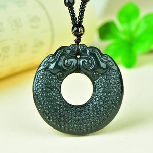 Puede incluir: Colgante de jade verde oscuro con un diseño circular y tallas intrincadas. El colgante está suspendido de un collar de cuentas negras. El jade tiene un acabado pulido y brillante, con un agujero central.