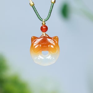 May include: A pendant necklace featuring a cat-shaped charm. The charm is a gradient of orange and white, with a heart-shaped cutout. The necklace has a green cord with gold accents and a red bead.
