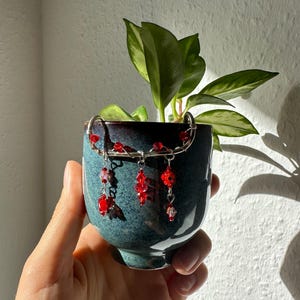 Könnte beinhalten: Kleiner, teal-farbener Keramiktopf mit silberfarbenem Drahtakzent und hängenden roten Kristallperlen. Der Topf enthält eine kleine grüne Pflanze mit bunten Blättern. Der Topf wird in einer Hand gehalten.