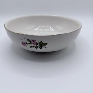 May include: A white ceramic bowl featuring a delicate floral pattern of pink roses and green leaves on the exterior side. The rim of the bowl is finished with a thin, metallic silver band that shows slight signs of wear and fading.
