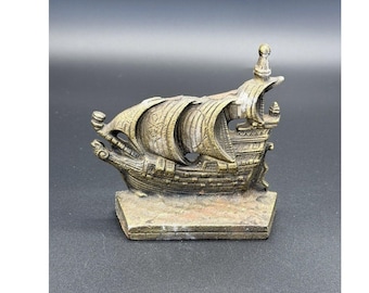 May include: A metallic bronze-toned ship figurine featuring detailed sails and a textured hull, mounted on a rectangular base. The sculpture displays a weathered finish with dark accents highlighting the intricate carvings on the vessel's structure.