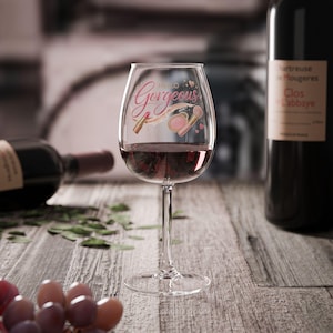 May include: A clear wine glass filled with red wine, featuring the text "Hello Gorgeous" in pink script, along with makeup illustrations. A bottle of red wine is in the background, and the glass sits on a wooden surface.