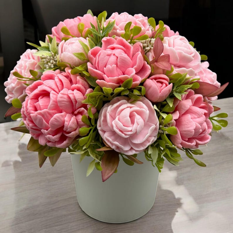 Pode incluir: Um buqu&ecirc; de flores de pe&ocirc;nia rosa em um vaso de cer&acirc;mica branco. As flores t&ecirc;m m&uacute;ltiplas camadas de p&eacute;talas e s&atilde;o cercadas por folhas verdes e pequenos bot&otilde;es. O arranjo est&aacute; em uma superf&iacute;cie cinza claro.