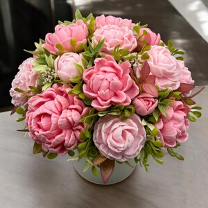 Pode incluir: Um buqu&ecirc; de flores de pe&ocirc;nia rosa com folhas verdes, arranjado em um vaso branco. As flores est&atilde;o em v&aacute;rios est&aacute;gios de flora&ccedil;&atilde;o, criando uma apar&ecirc;ncia cheia e texturizada. O arranjo est&aacute; em um fundo neutro.
